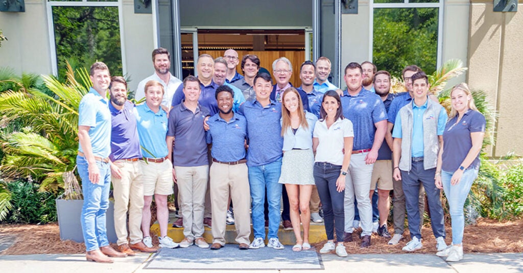 A group of 20 people stand smiling together outside a building entrance, dressed in casual and business-casual attire, with greenery and sunny weather in the background.