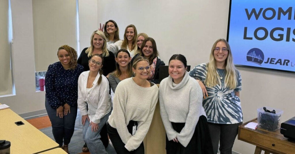 A group of twelve women pose and smile in a conference room. Some stand, others kneel in front. A screen in the background displays partial text reading WOM... LOGIS.... The atmosphere is casual and friendly.