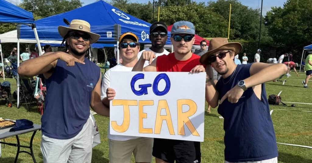Five men stand outdoors on grass, smiling and pointing at a large white sign that says GO JEAR in blue and orange letters. Tents and people are visible in the background under a sunny sky.