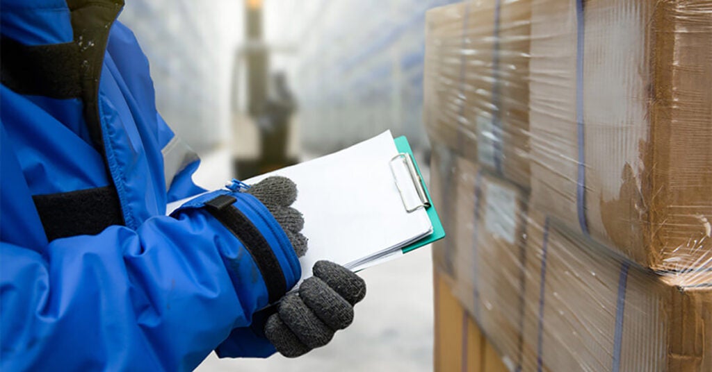 A person in a blue jacket and gloves holds a clipboard and pen, checking stacked boxes wrapped in plastic in a warehouse or storage facility.