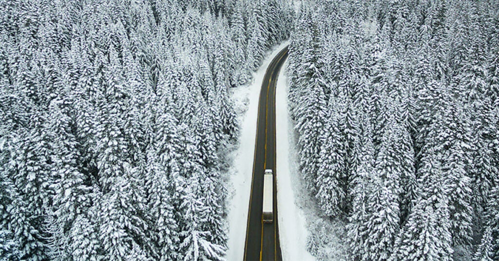 A truck drives along a winding road surrounded by dense, snow-covered pine trees in a wintry forest landscape.