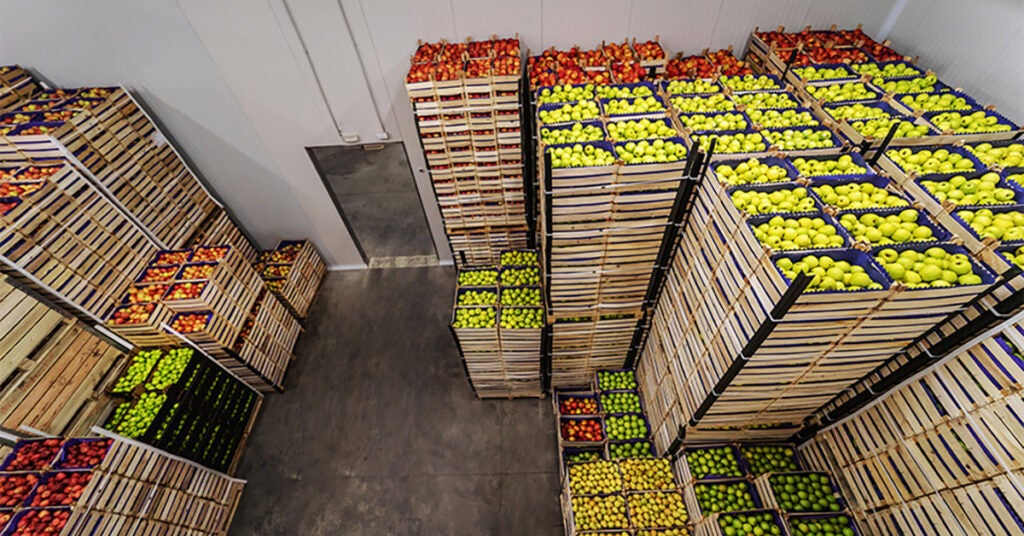 Stacks of wooden crates filled with red, yellow, and green apples are organized neatly in a large warehouse with concrete floors and white walls.
