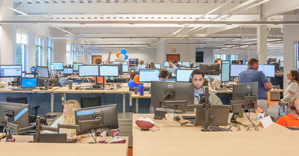 A modern open-plan office with people working at desks, multiple computer monitors on each desk, bright lighting, and a few blue and orange balloons in the background—ideal for businesses seeking comprehensive freeze protection service.