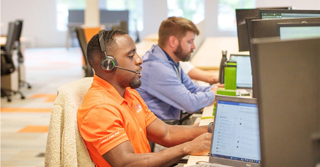 Two men work at computers in an office. The man in the foreground, wearing an orange shirt and headset, speaks into his microphone. The man in the background, in blue, types at his desk. Multiple monitors are visible.
