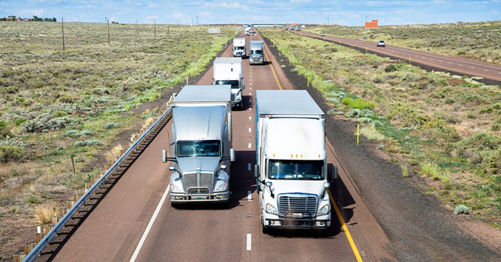 Several large semi-trucks and vehicles travel on a divided highway through a grassy, open landscape under a partly cloudy sky, where freeze protect measures are essential for safe and reliable transport.