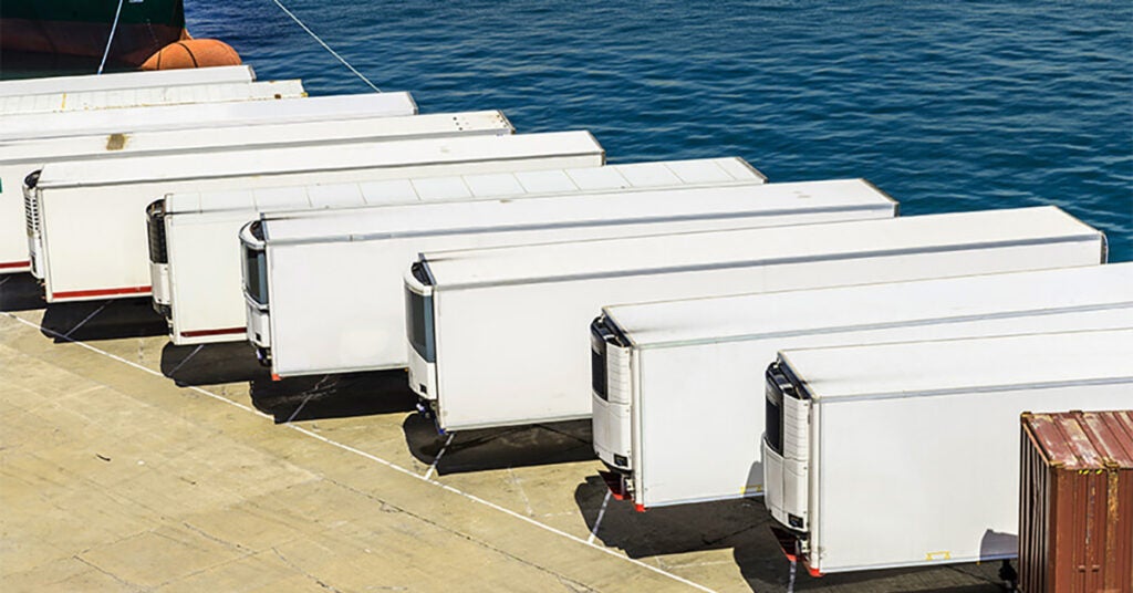 Several white refrigerated semi-trailers are lined up side by side on a concrete dock next to a body of water, with a cargo container nearby.