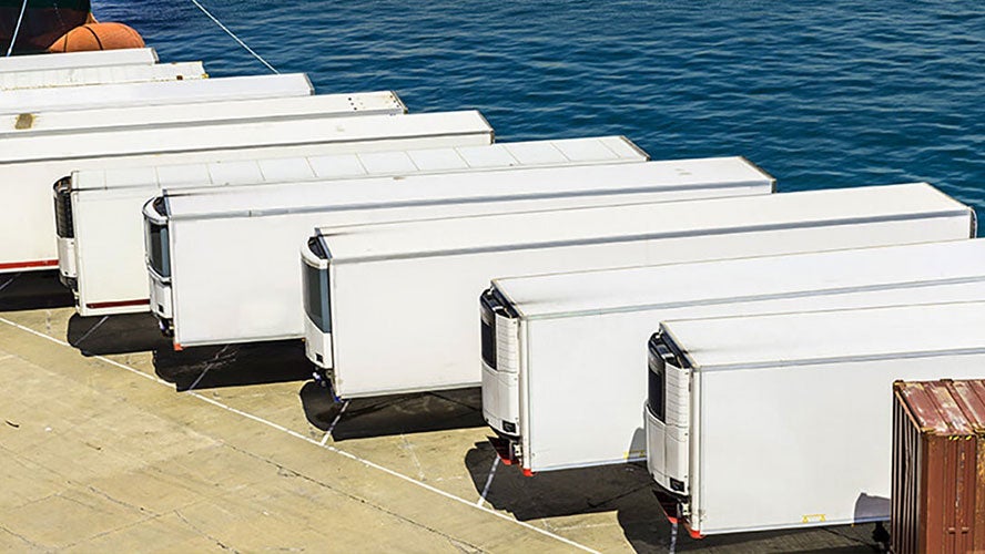 Several white semi-truck trailers, ideal for temperature-controlled shipping, are parked in a row on a concrete dock beside blue water. A brown shipping container sits in the lower right corner.
