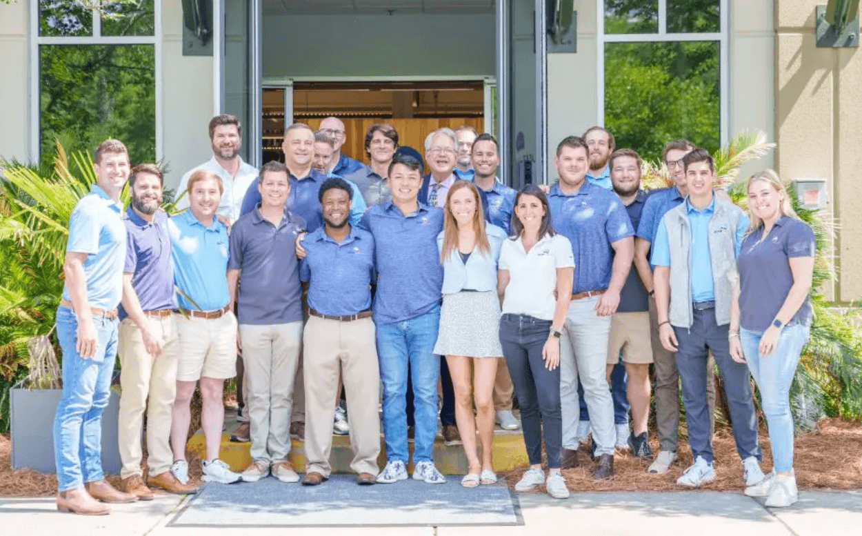 A group of 20 people, mostly men and a few women, stand smiling together outside a building entrance on a sunny day, dressed in casual business attire with light blue tones—ideal for those seeking sales jobs in logistics. Lush green plants surround the entrance.