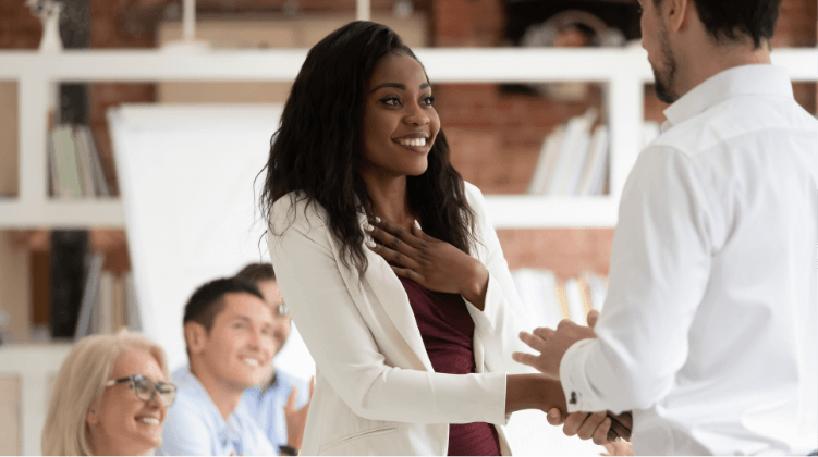 A smiling woman in a white blazer shakes hands with a man while colleagues sit in the background, watching and clapping—a great moment for anyone pursuing logistics sales career opportunities in a bright office setting.