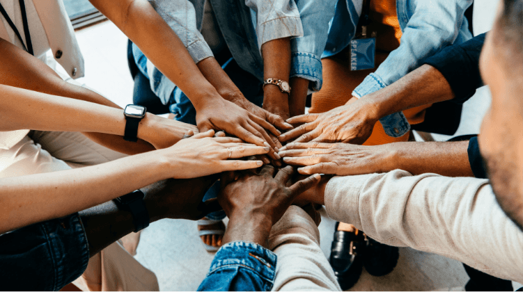 A group of diverse people sit in a circle and stack their hands together in the center, symbolizing teamwork, unity, and collaboration—key values for anyone pursuing a logistics sales career.