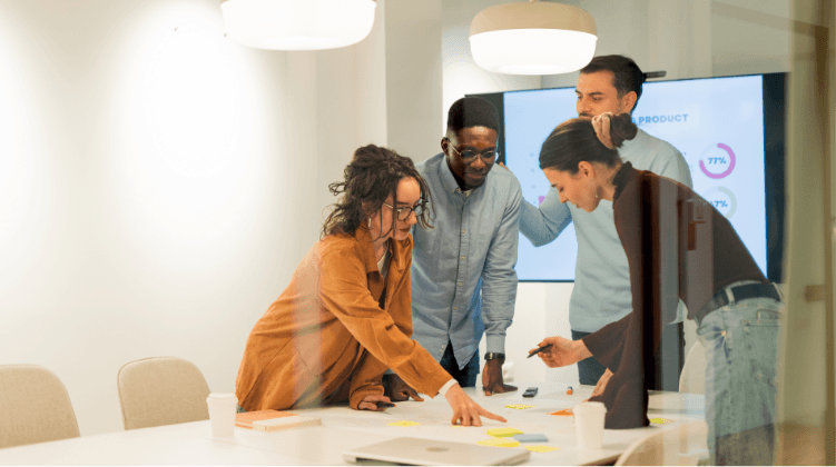 Four people in an office are gathered around a table with sticky notes, discussing ideas for a logistics sales career. A screen in the background displays charts. The atmosphere appears collaborative and focused.