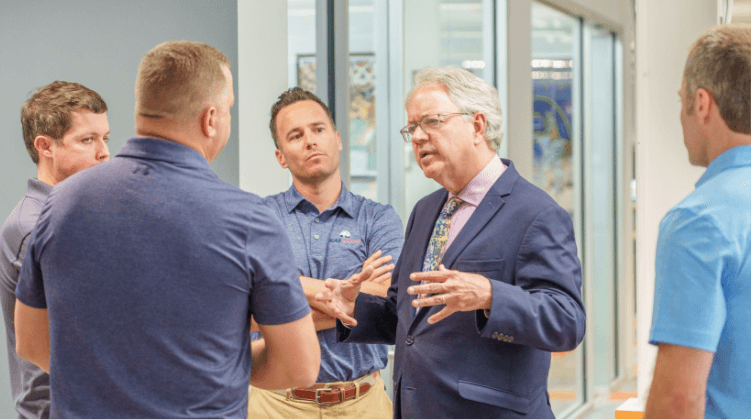 Five men stand in a modern office, engaged in discussion. Four wear collared shirts, while one older man in a suit and tie gestures as he speaks—perhaps sharing insights about sales jobs in logistics or advancing a logistics sales career.