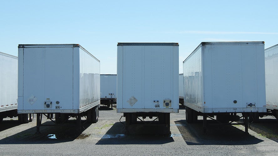 Three white, empty semi-trailers—ideal for temperature-controlled shipping—are parked side by side in a gravel lot under a clear blue sky. The image is taken from directly behind the trailers.