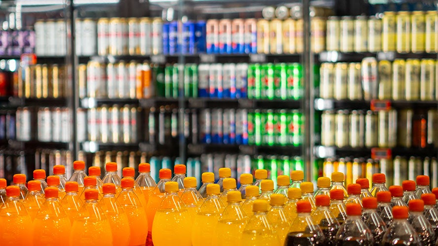 Rows of plastic bottles filled with orange and yellow drinks are displayed in front of a refrigerated section, showcasing the variety within the food and beverage supply chain. The blurred background keeps focus on the vibrant drinks in the foreground.