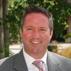 A man in a gray pinstripe suit and pink striped tie smiles outdoors, with blurred greenery and sunlight in the background—reflecting the professionalism found in full-service third-party logistics and temperature-controlled 3PL shipping.